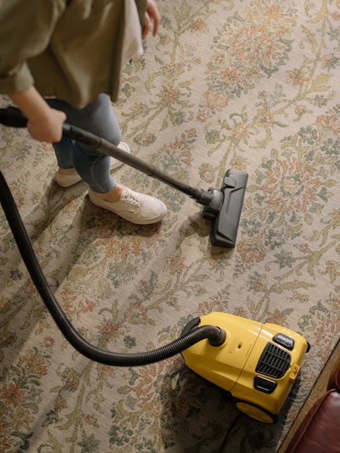 Close-up of a person operating a black vacuum cleaner on a patterned area rug in a well-lit living room. The rug features intricate floral designs in beige, green, and orange tones. The vacuum cleaner