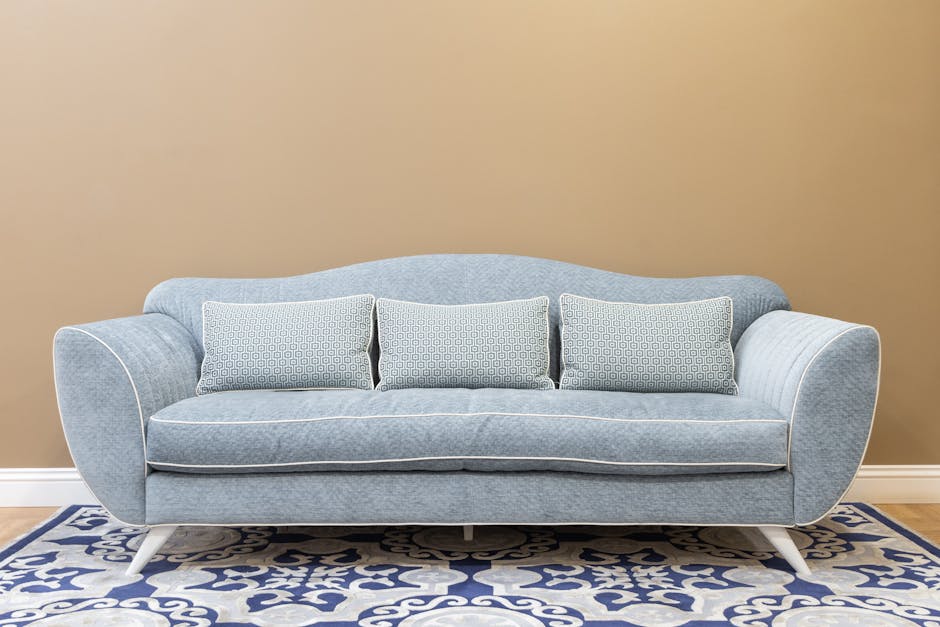 A light blue upholstered sofa with rolled arms and three patterned cushions, positioned against a beige wall in a living room. The sofa has a clean, deep-cleaned appearance, indicating recent deep cle