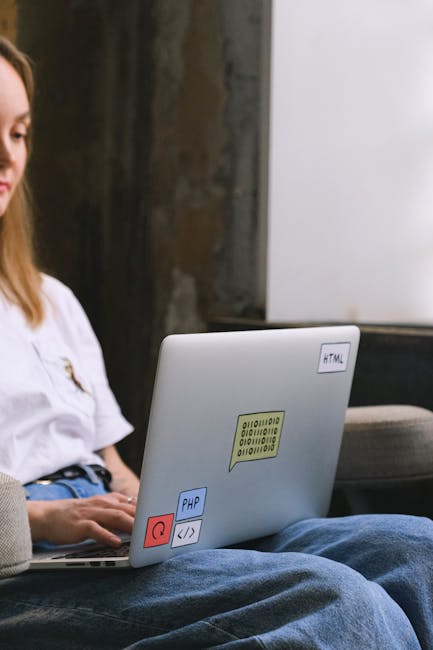 A close-up of a silver laptop resting on a dark leather armchair in a modern office setting. The laptop's surface features colorful stickers, including a yellow binary code pattern, a blue 'PHP' label